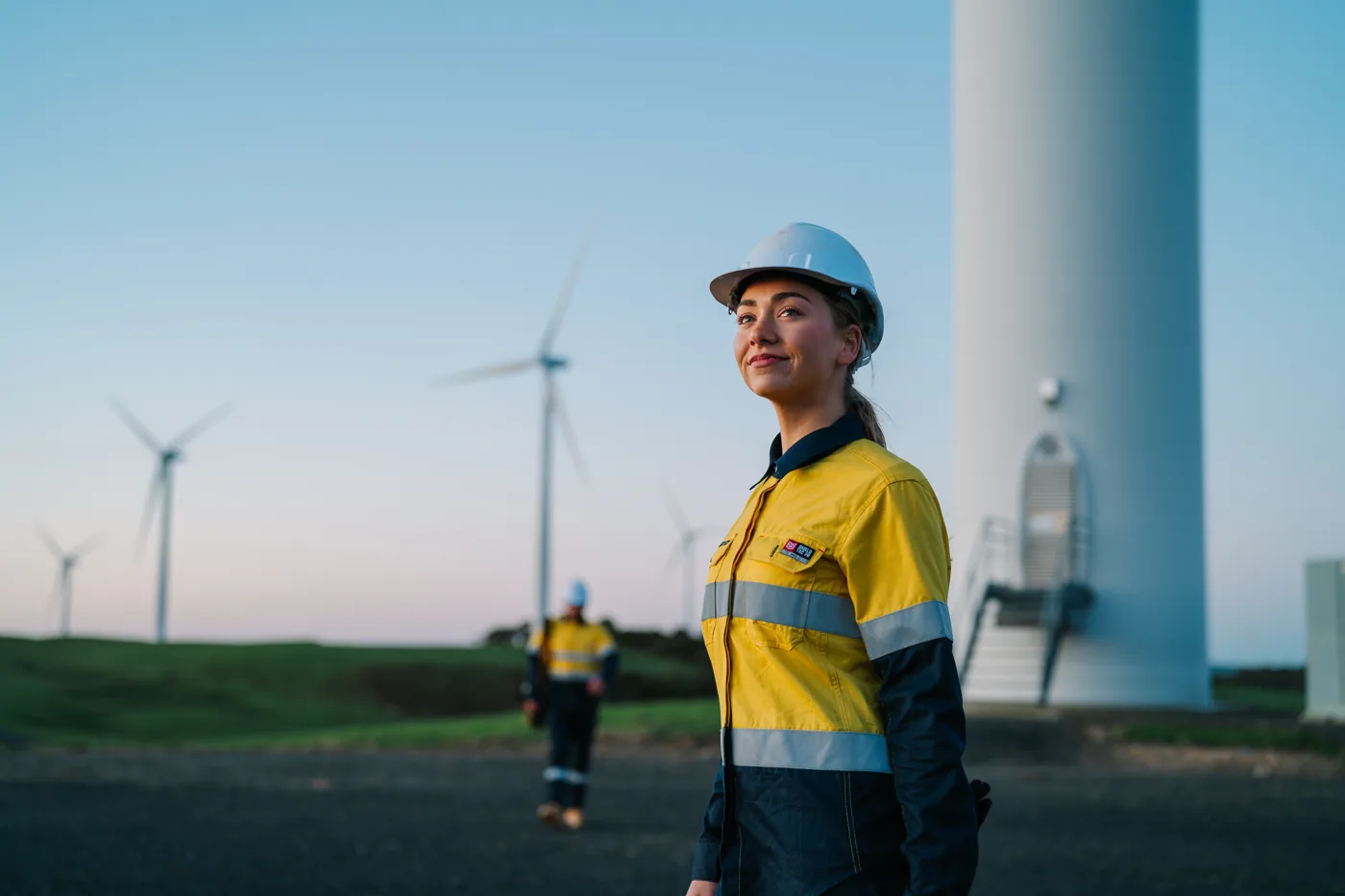 A woman in a hard hat and hi-vis KingGee workwear standing in front of wind turbines at dusk.
